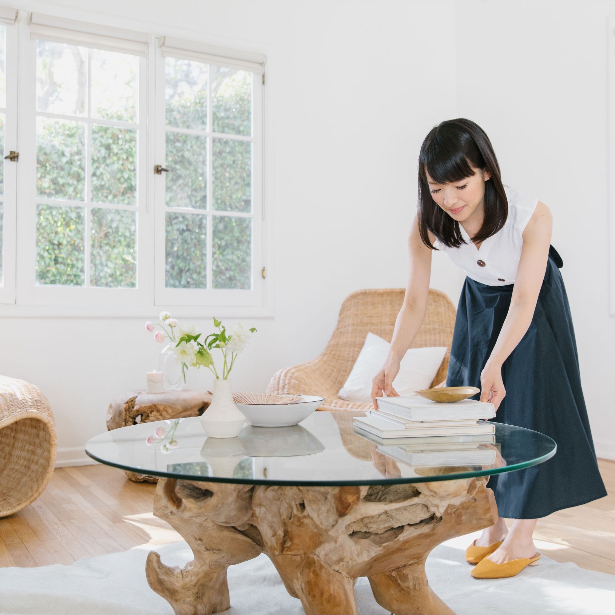 Tidying guru Marie Kondo wears a white top and a blue skirt, and tidies books sitting on top of a white coffee table with natural wooden legs