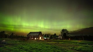 wispy lines of green and yellow light stretch across the sky above a barn and a treeline