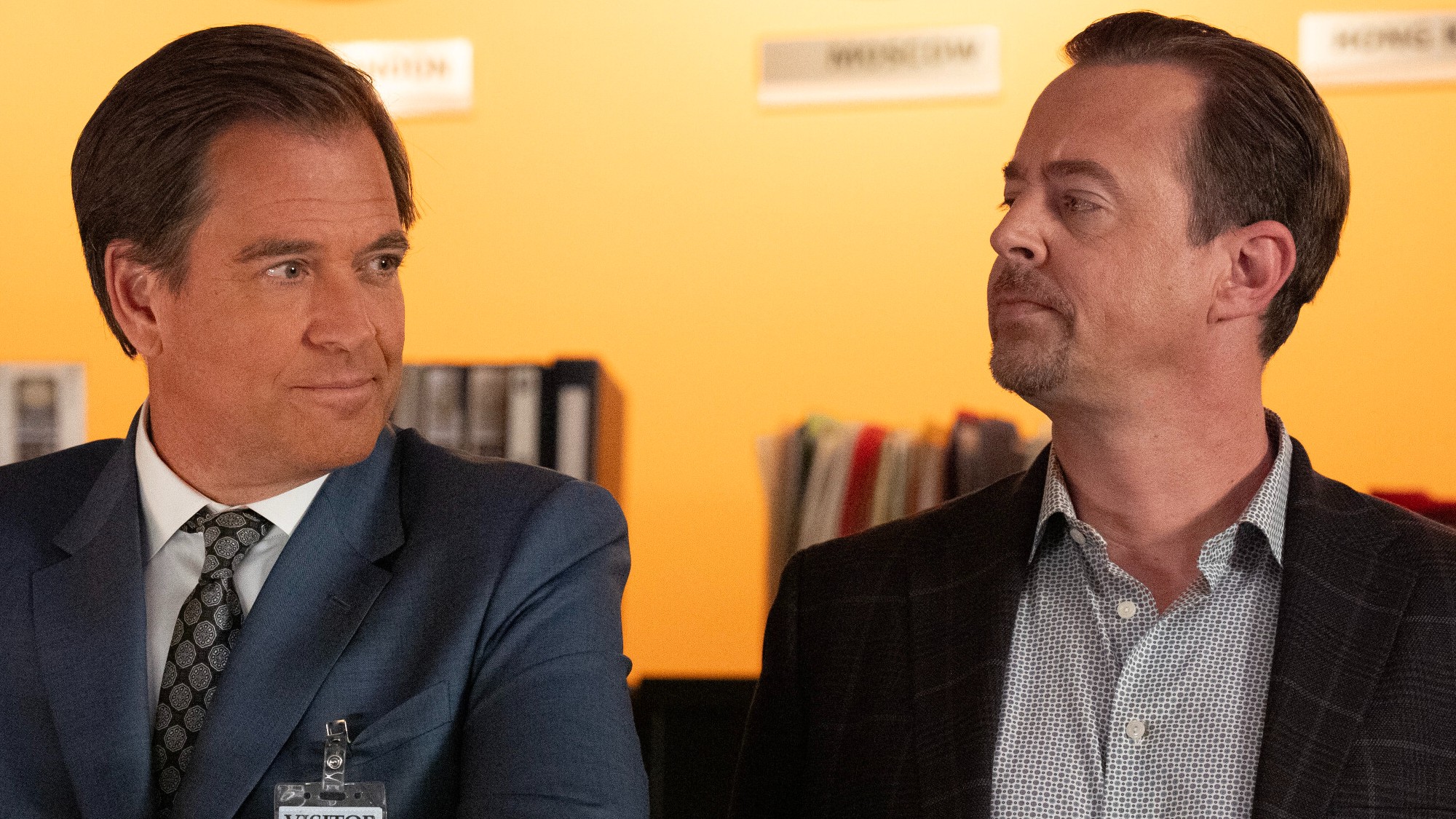 Tony and McGee lock eyes as they stand side-by-side in the NCIS office. They're both wearing sport coats and collared shirts. 