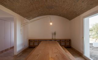 Interior view of a space at Casa Modesta featuring white walls, tiled floor, arched brick ceiling, wall lamp, wooden dining table with bench seats, wooden shelving unit, two plants and a large glass door