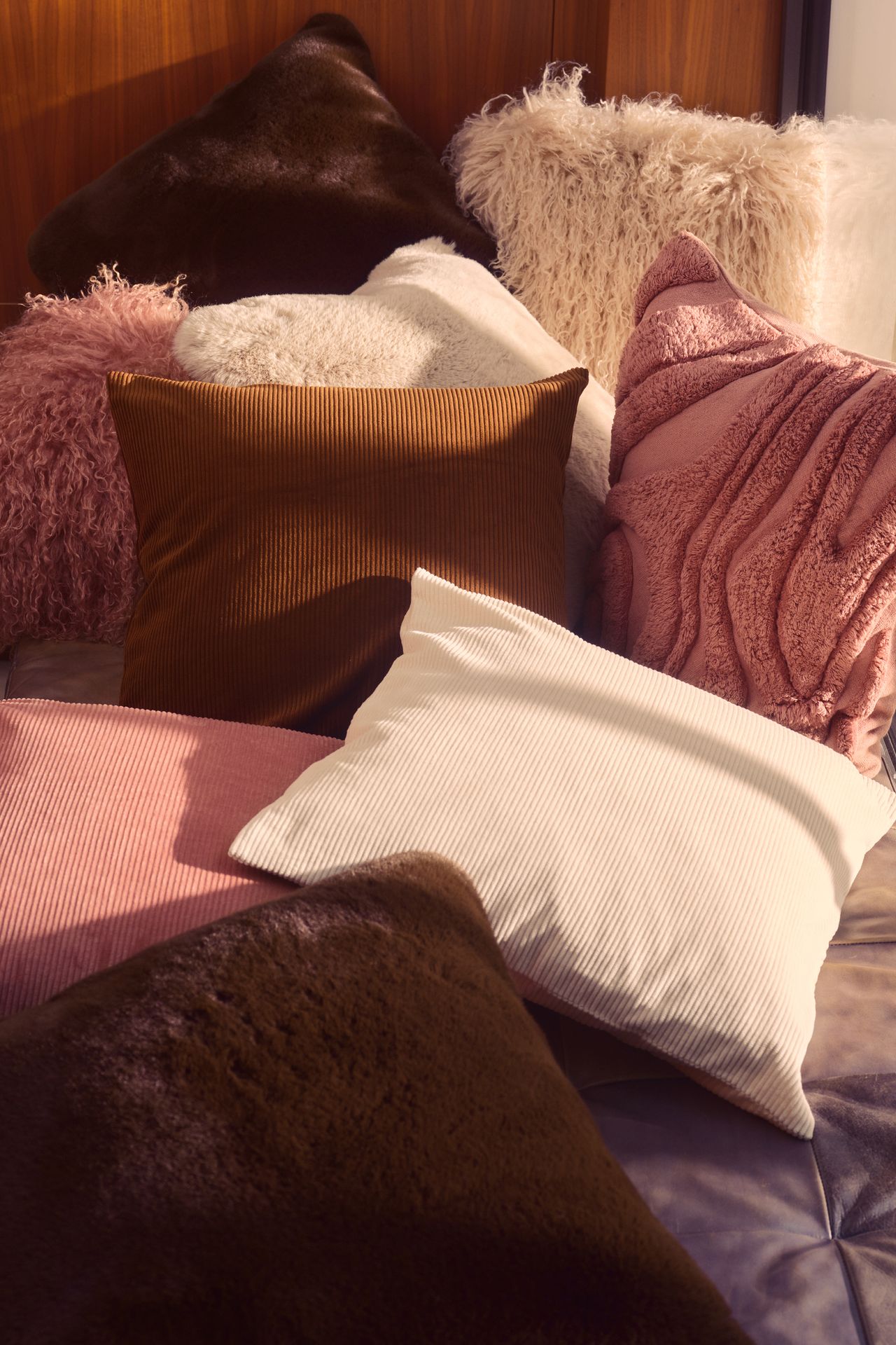 Image of a pile of different pillows that are either brown, white, or rose pink. They are also different textures as well &mdash; some are shag, some are boucle, and some are corduroy.