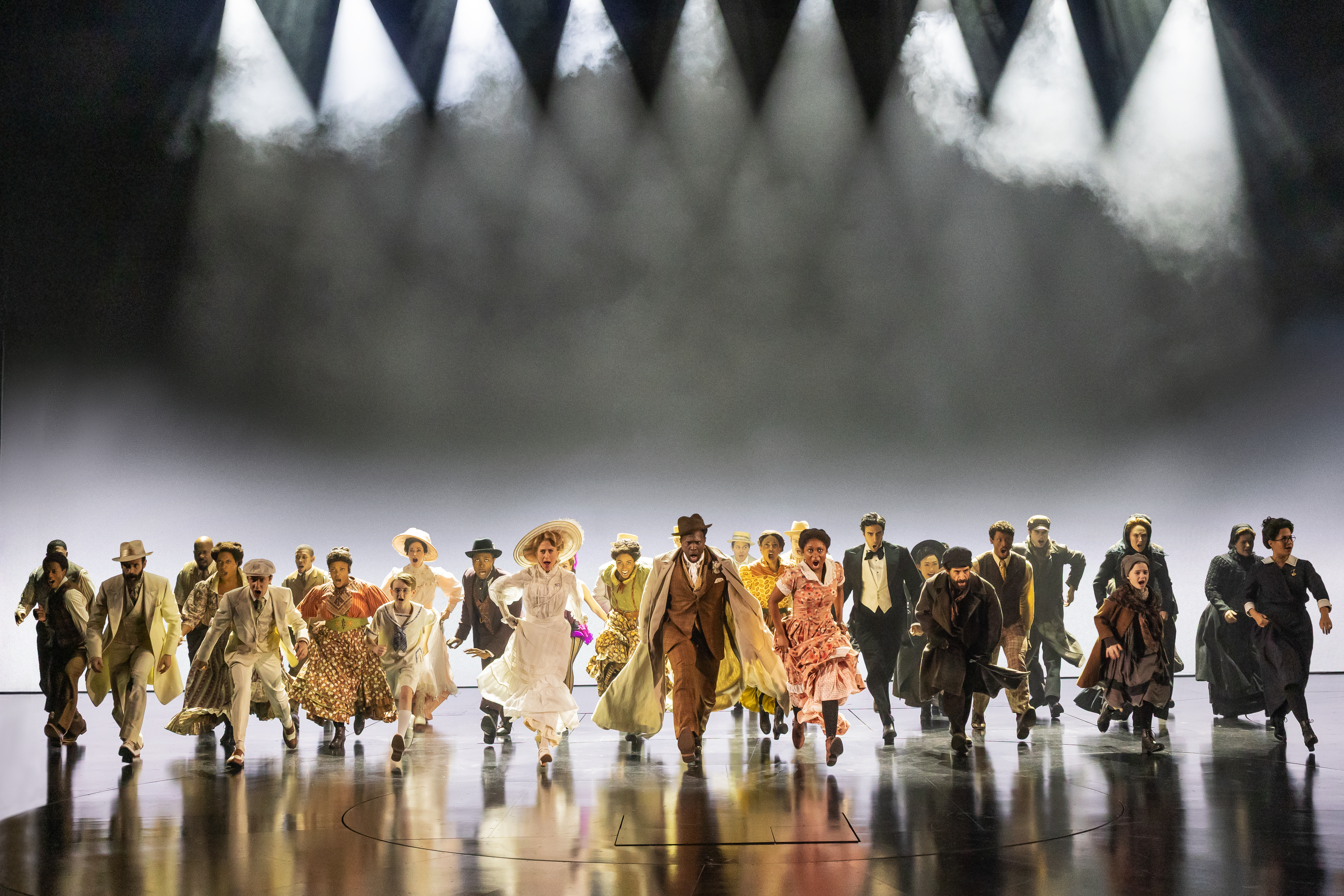 the cast of ragtime dressed in 20th century clothing standing in a line on a dark stage in the broadway musical revival of ragtime