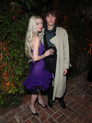 Anya Taylor-Joy has long blonde hair and wears a purple dress with black pumps while her husband Malcolm McRae wears a tan trench coat