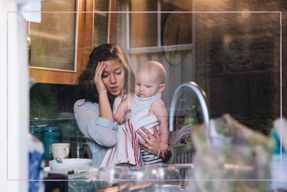 A mum looking stressed while holding a baby, pictured through a kitchen window