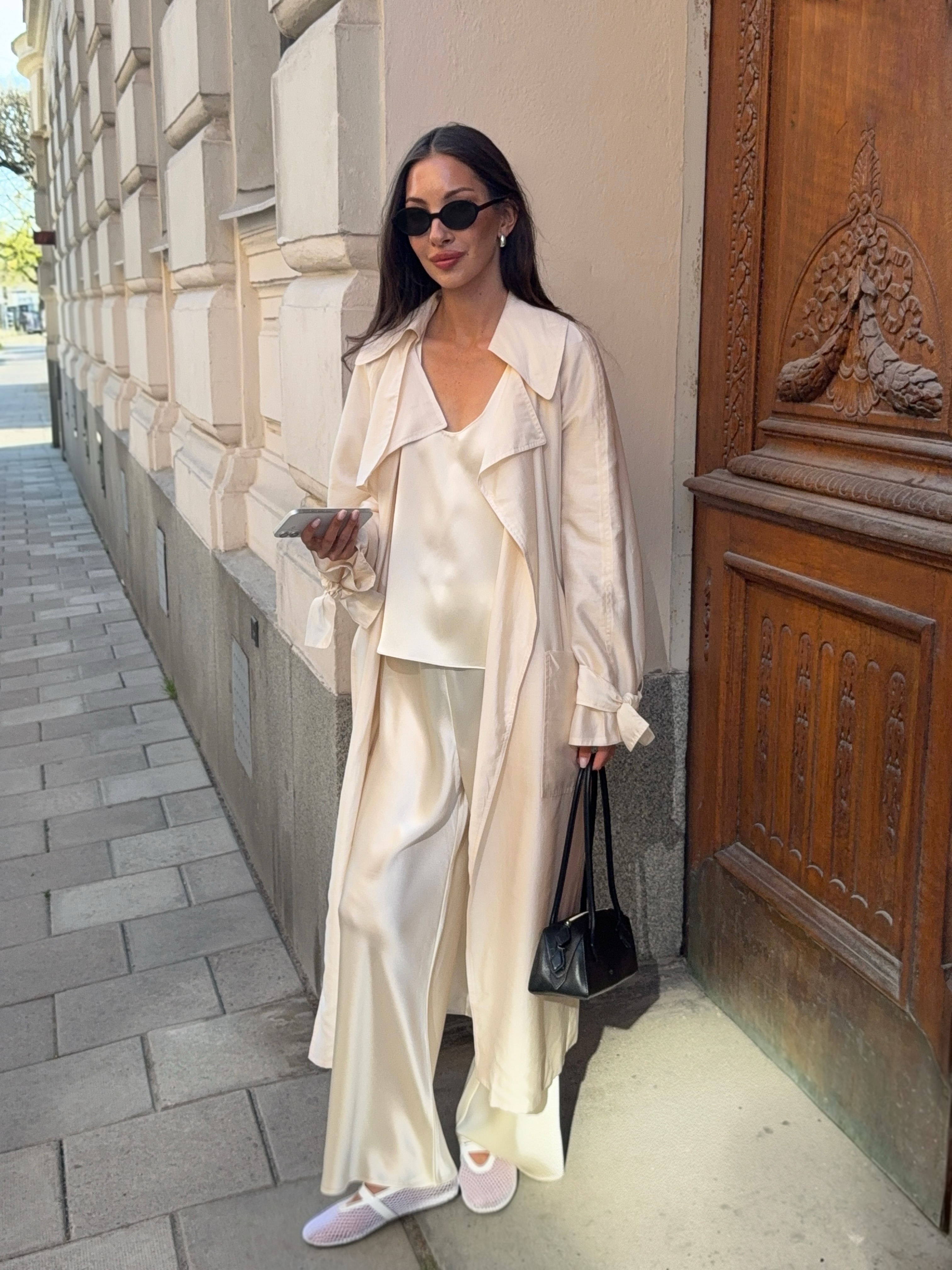 An image of an influencer wearing a monochromatic cream-coloured outfit, featuring a satin trench coat, satin top and satin trousers, along with a black handbag and white mesh flats