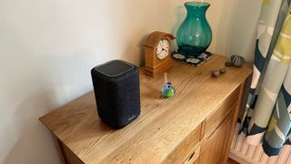 Denon Wireless Speaker 150 on a wooden side table with a clock, a glass vase and glass bird