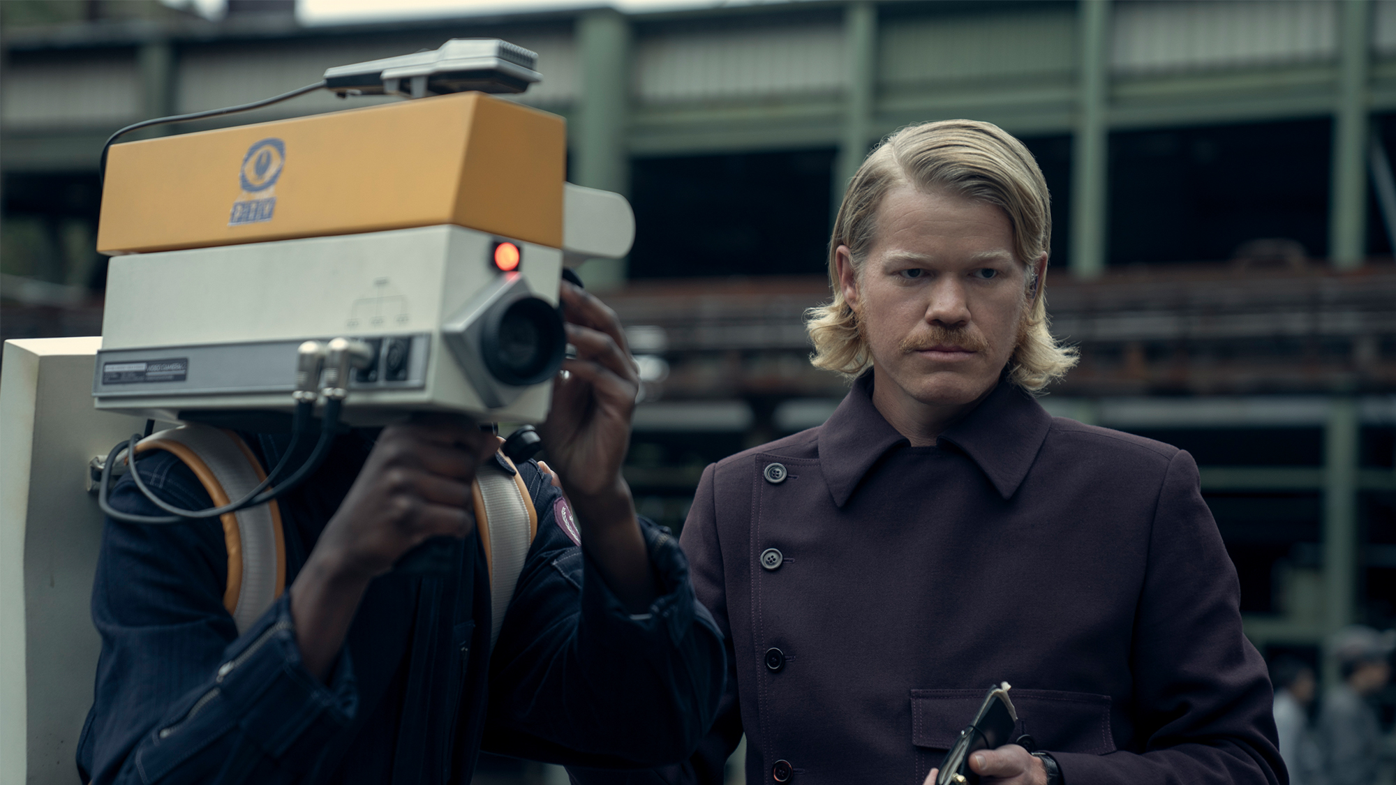 Jesse Plemons in a purple jacket, standing next to a man holding a tv camera in Sunrise on the Reaping
