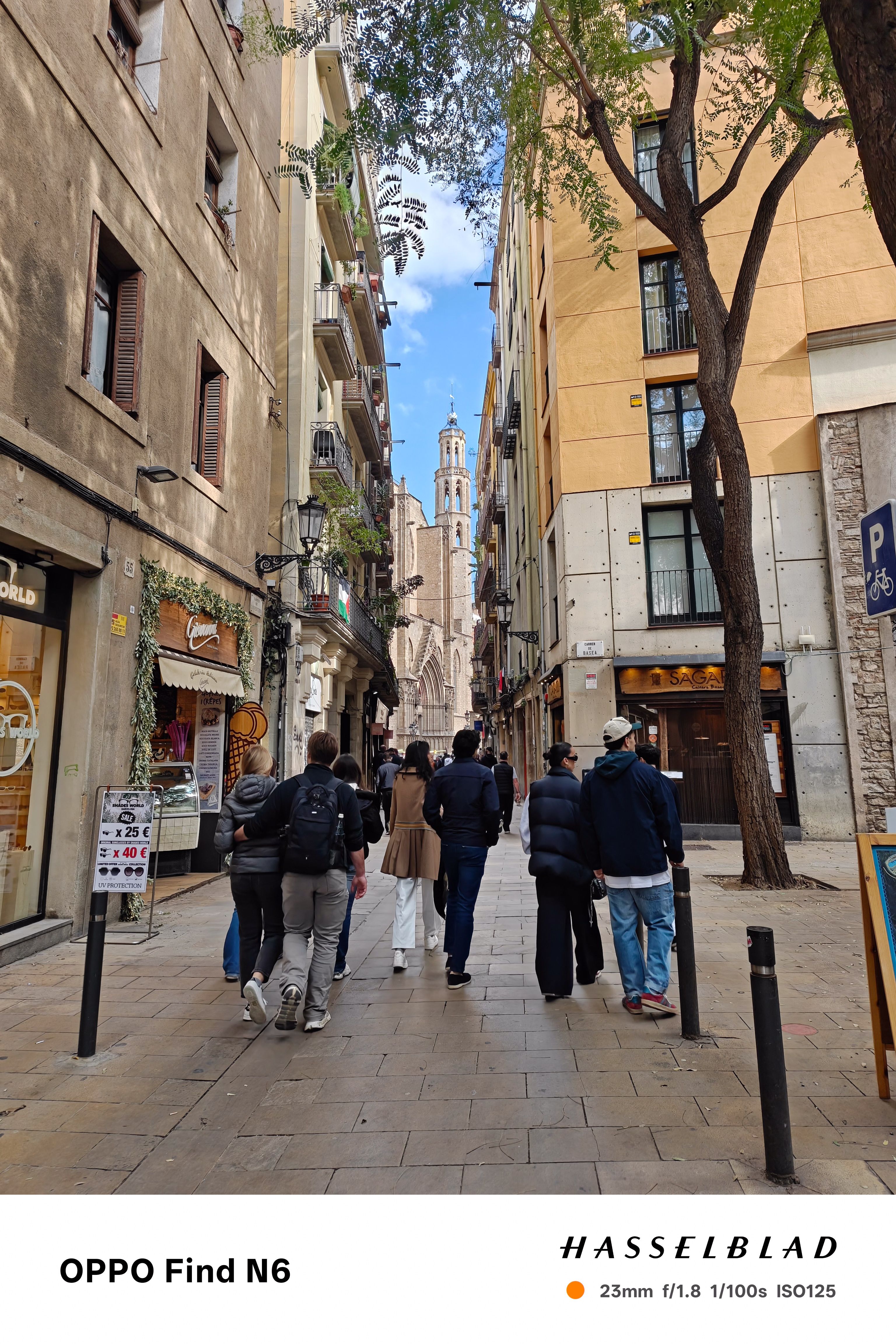A wide-angle street photograph of a narrow, historic European alleyway. Several people are walking away from the camera toward a tall, slender cathedral spire in the distance. The scene is framed by tall, warm-toned buildings and a large tree on the right.