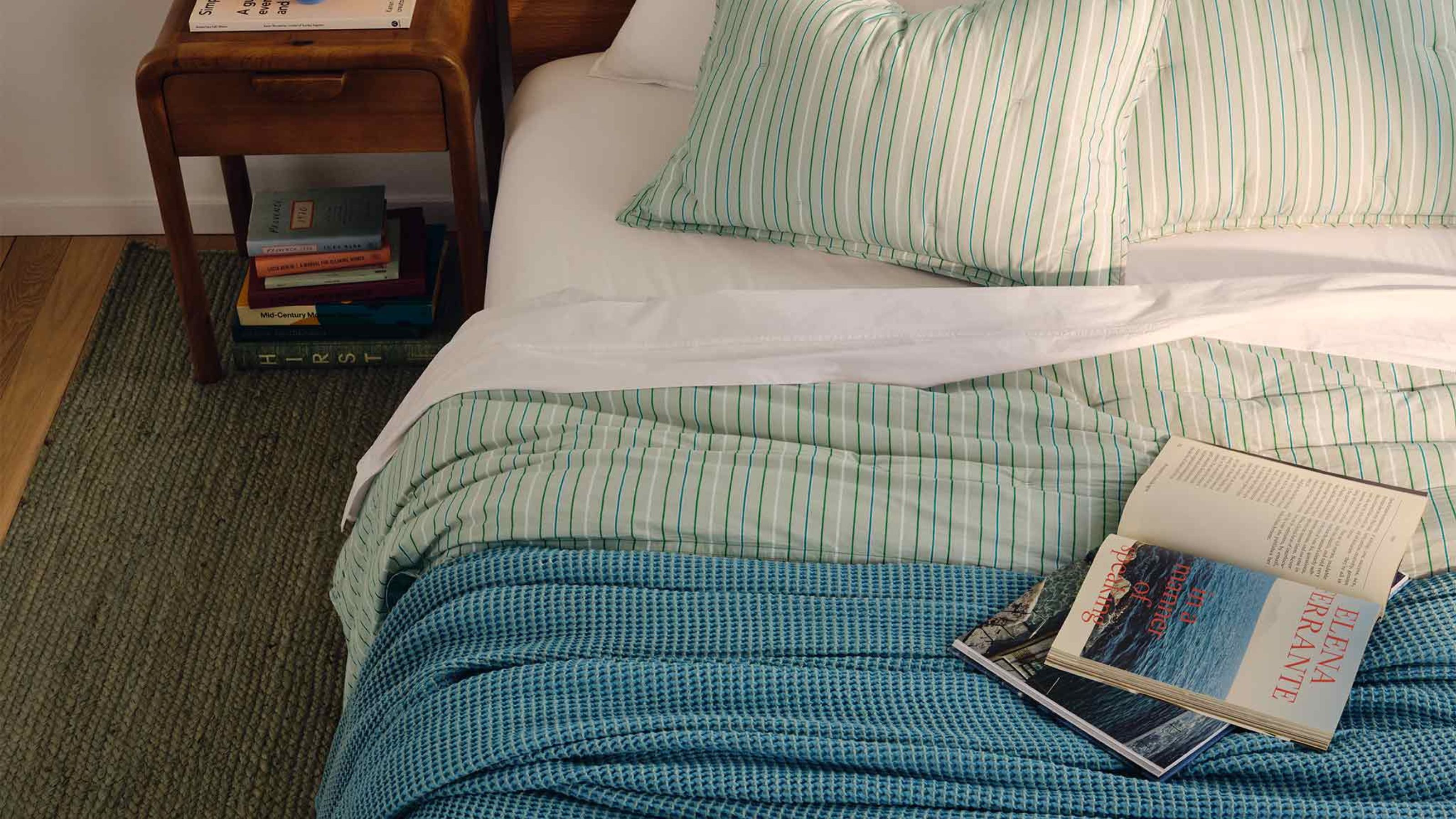 Brooklinen blue cotton waffle blanket on a double bed with striped green bedding with an open book beside a dark wood nightstand with a stack of books.
