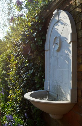 an athenian wall fountain in the middle of a lush green hedge