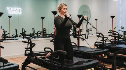 Woman exercising on Lagree Megaformer machine. She kneels on the platform and holds D handles connected to cables in front of her with elbows bent