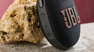 A closeup of the control buttons on the side of the JBL Clip 5, pictured leaning against a sand coloured rock against a pink background,