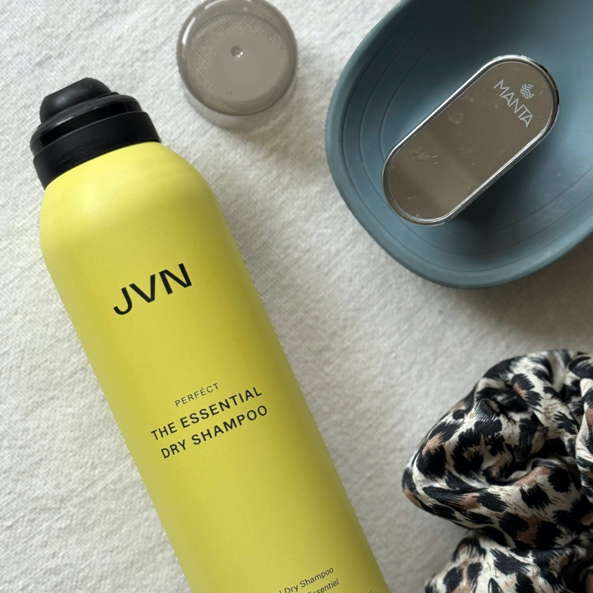 Bird's eye view of JVN Hair The Essential Dry Shampoo without its lid, a slate blue Manta Hair Brush and a leopard print scrunchie, set on an off-white linen background