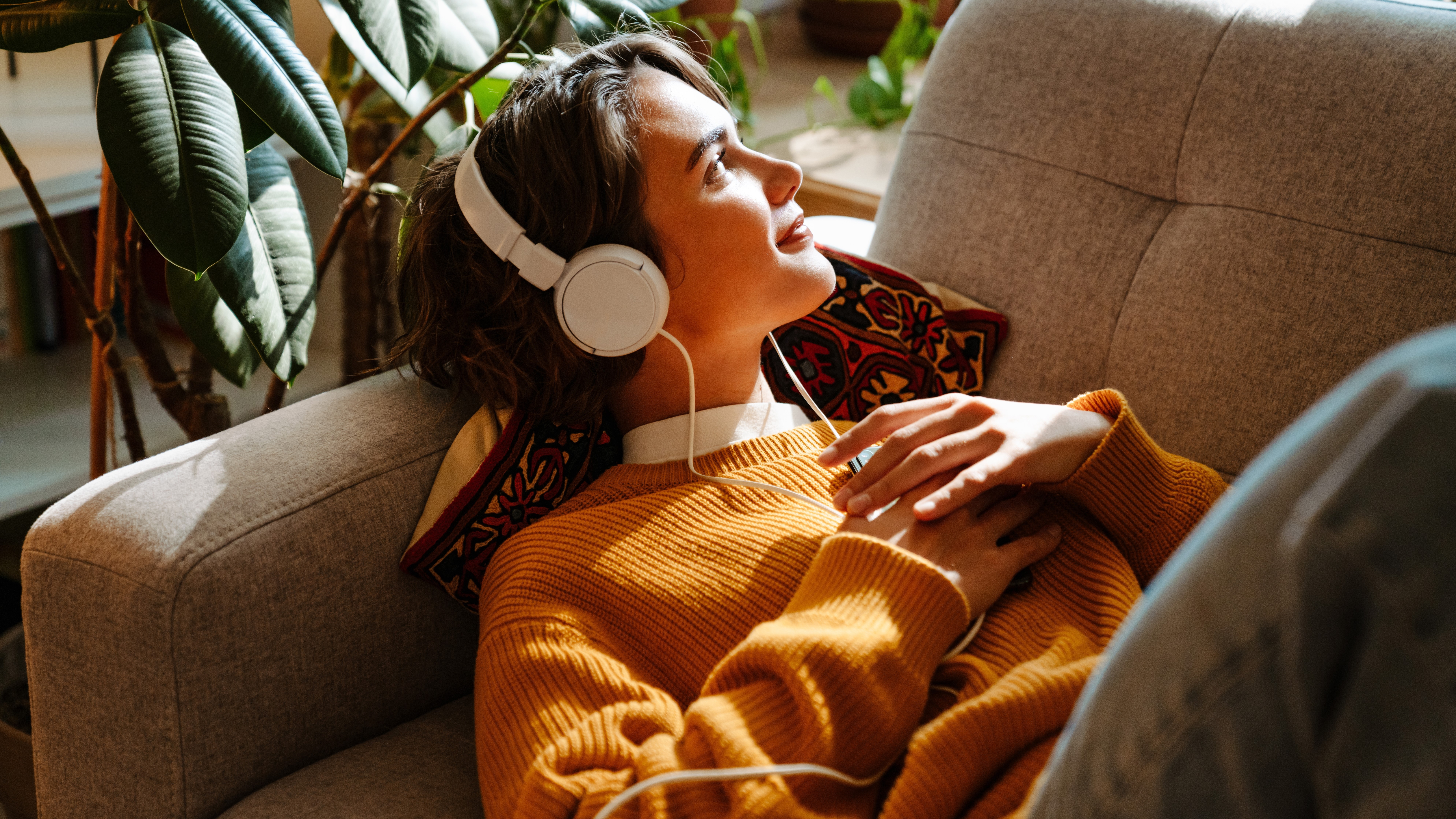 Mujer escuchando música con sus auriculares mientras descansa en el sofá y sostiene su teléfono y mira a lo lejos