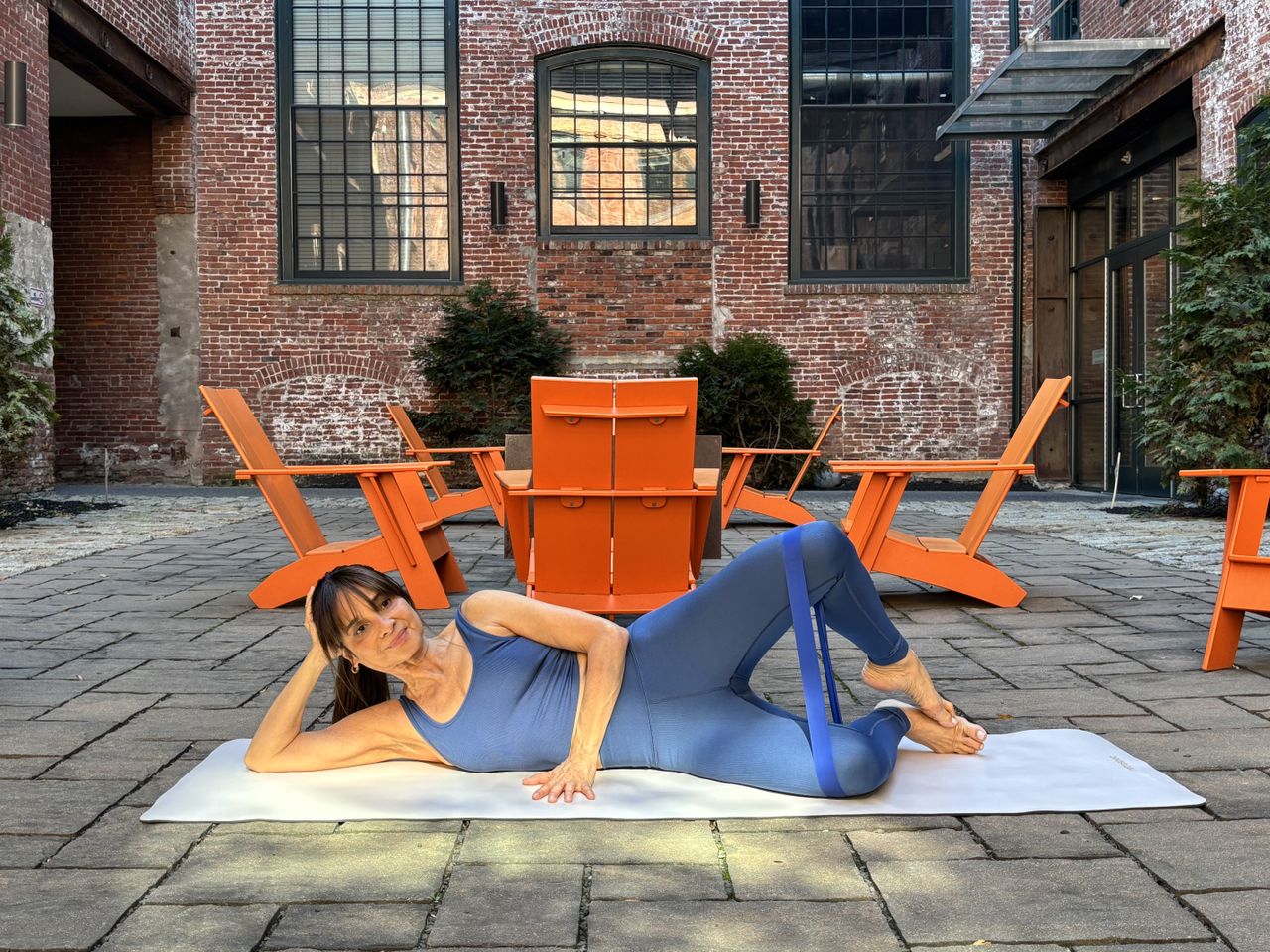 Pilates teacher Monica Borodin performs clamshell exercises. She wears a vest and leggings and exercises on a mat outside. Behind her we see a large brick building with long windows and a tablet and chairs in a courtyard. She is lying on her side with her legs bent and open, so that her feet are touching and knees wide apart. One leg lies on the floor while the other is pointing upwards. There is a resistance band looped around her thighs.