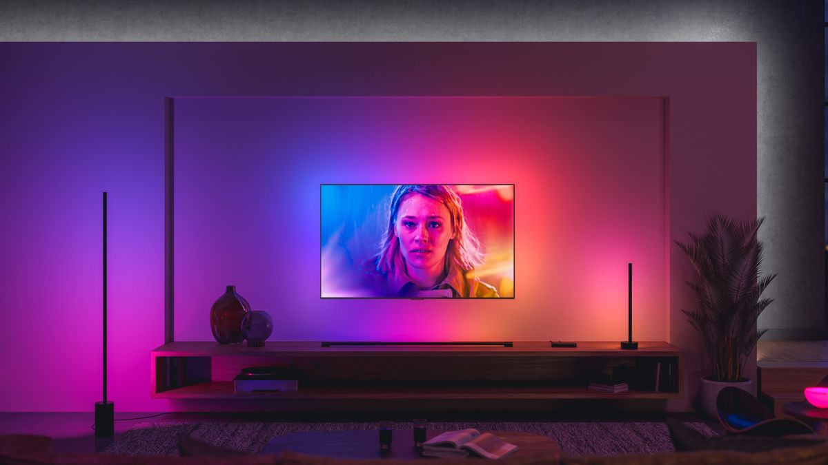 A modern living room with glowing Philips Hue lights, a wall mounted TV with a woman on screen and a wooden media unit below.