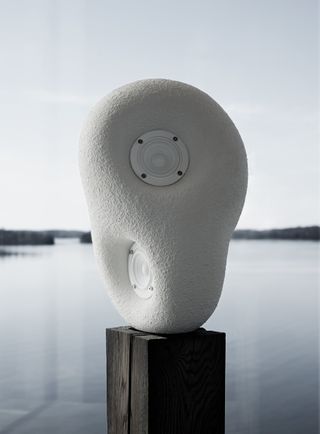 Light grey pebble-shaped ‘Acoustic Sculpture’ speaker on a wooden plinth with lake in the background