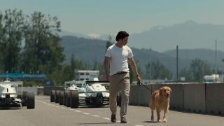 A man and a dog walk on a race track in front of race cars.