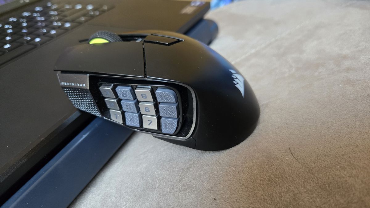 A Corsair Scimitar Elite Wireless SE gaming mouse on a carpet next to a gaming laptop and Stream Deck.