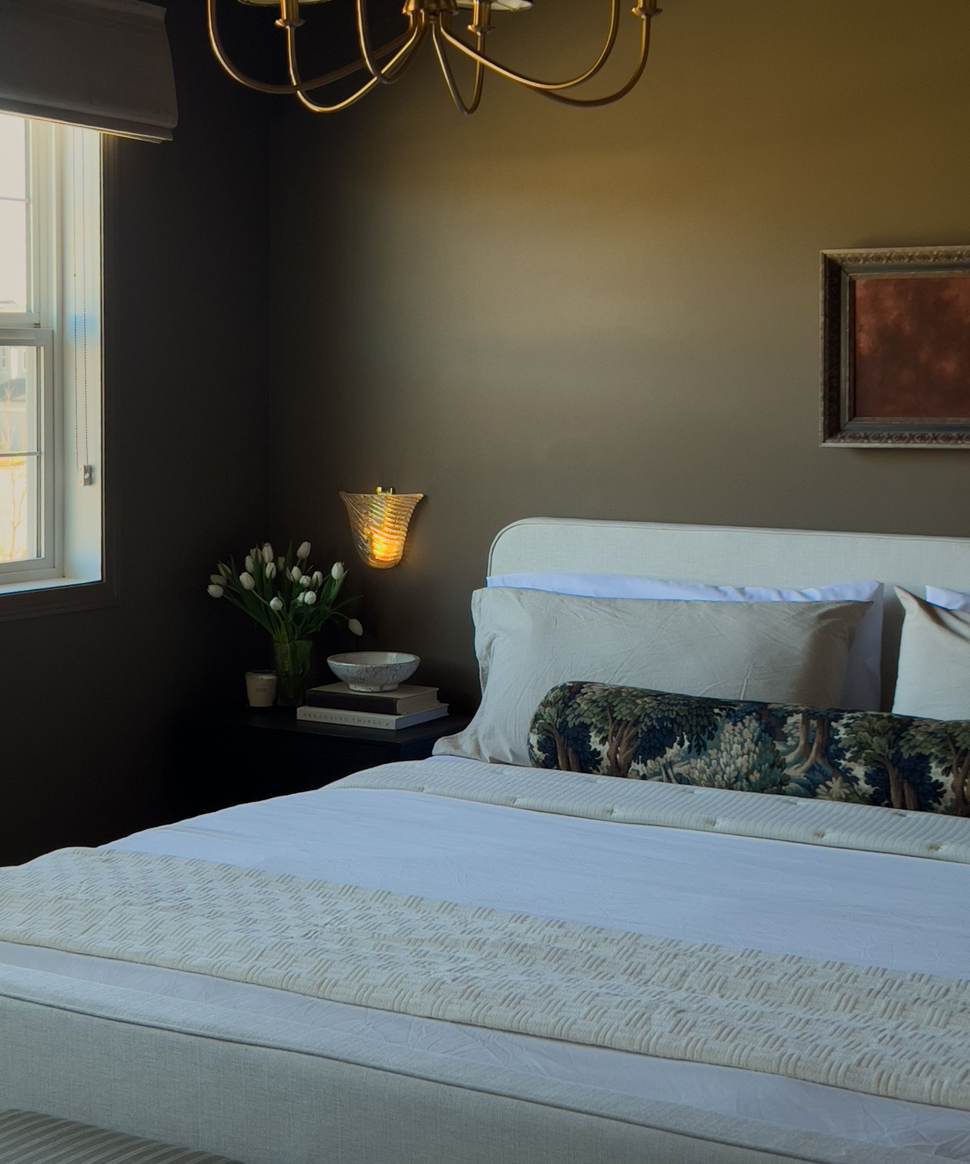 Bedroom with broccoli-brown painted walls, white bed with quilted white bedding, a jacquard bolster pillow, and a nightstand with a glass sconce above it and a vase of tulips.