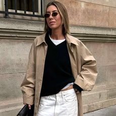 stylist and fashion influencer Hannah Lewis poses on a sidewalk wearing small oval sunglasses, a trench coat, a white T-shirt, black V-neck sweater, and white jeans