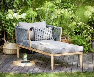 An outdoor lounger chair with grey cushioned decor and wooden frame on grey decking with wicker basket decor