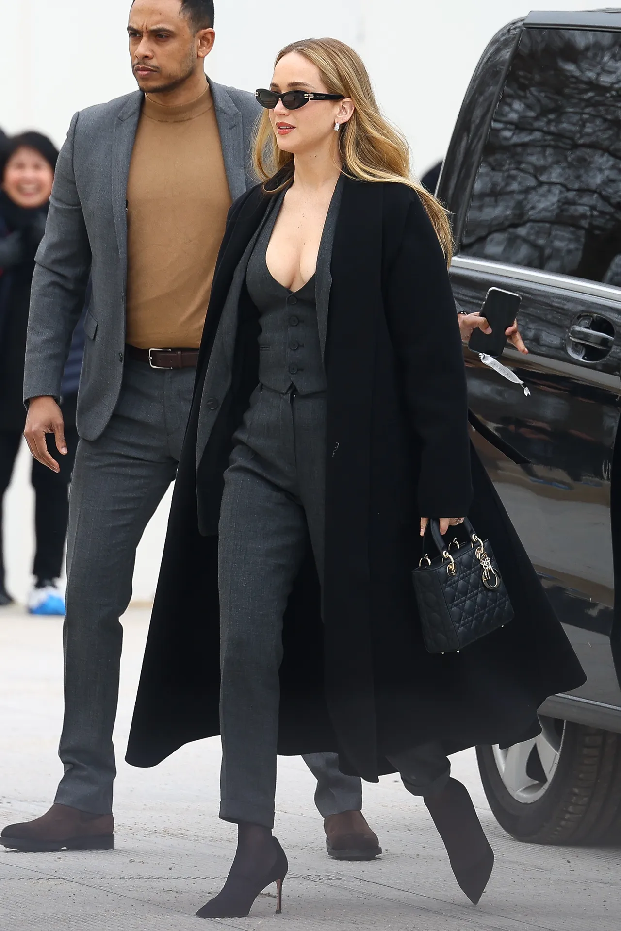 Jennifer Lawrence wearing a gray tailored waistcoat, blazer, and skinny pants with pumps and a black coat at the Dior show in Paris.