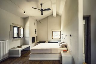 A double bed with concrete base and 2 pillows with 2 black lamps hanging from the wall on both sides of the bed. The room has cream walls and ceiling with brown wood flooring.
