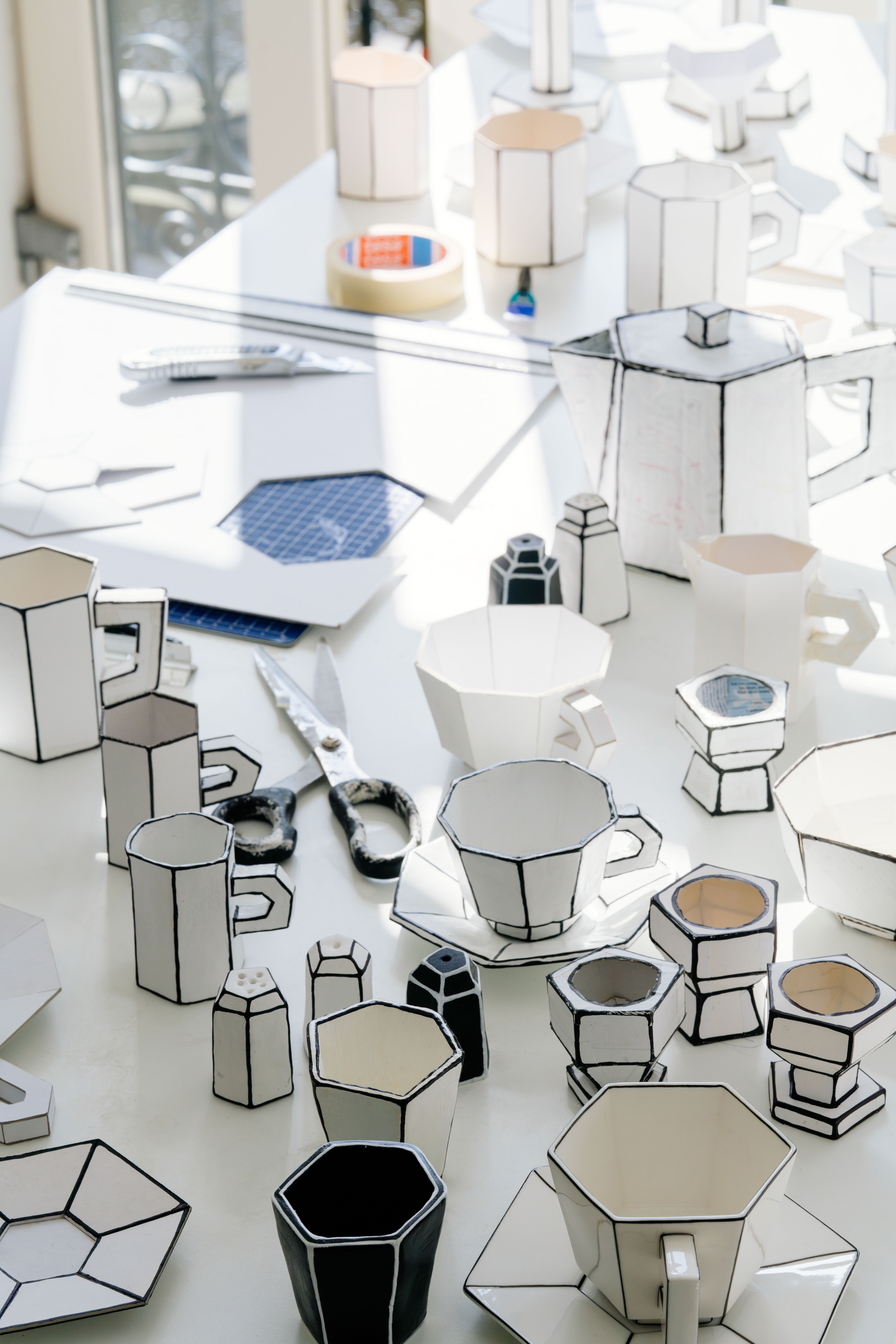 Cardboard mugs and ceramic ones inspired by them sitting on a white table in the sun.