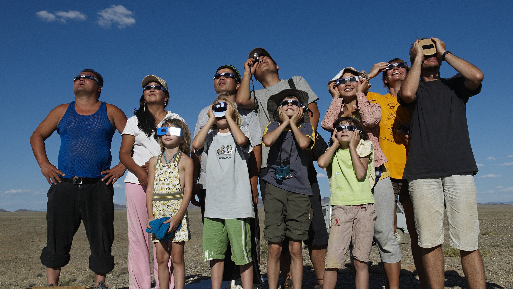 a group of people watch a solar eclipse wearing eclipse glasses and look up at the sun smiling.