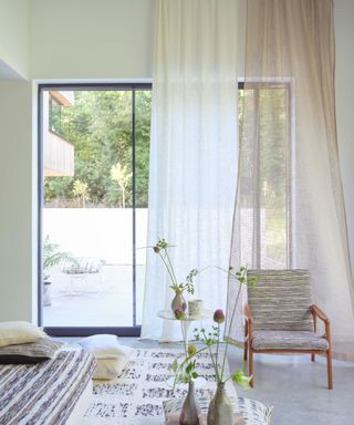 A light and airy decluttered bedroom in neutral colors with a sunny outdoor area visible through floor to ceiling windows and voile panels