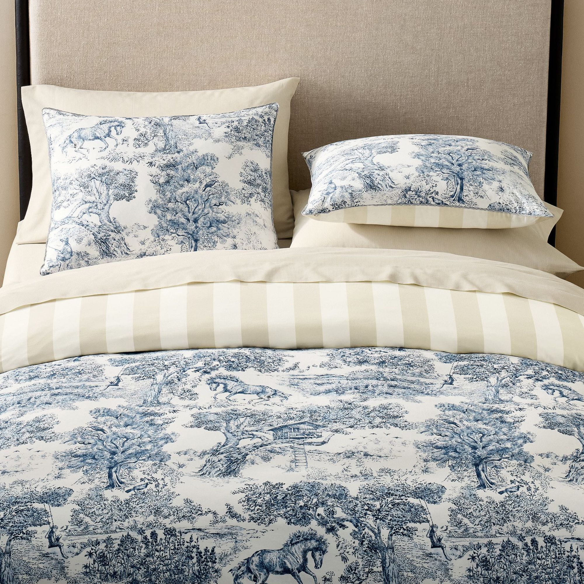 Taupe fabric bedframe with blue and white toile bedding and pillows with a reversible cream and white stripe design