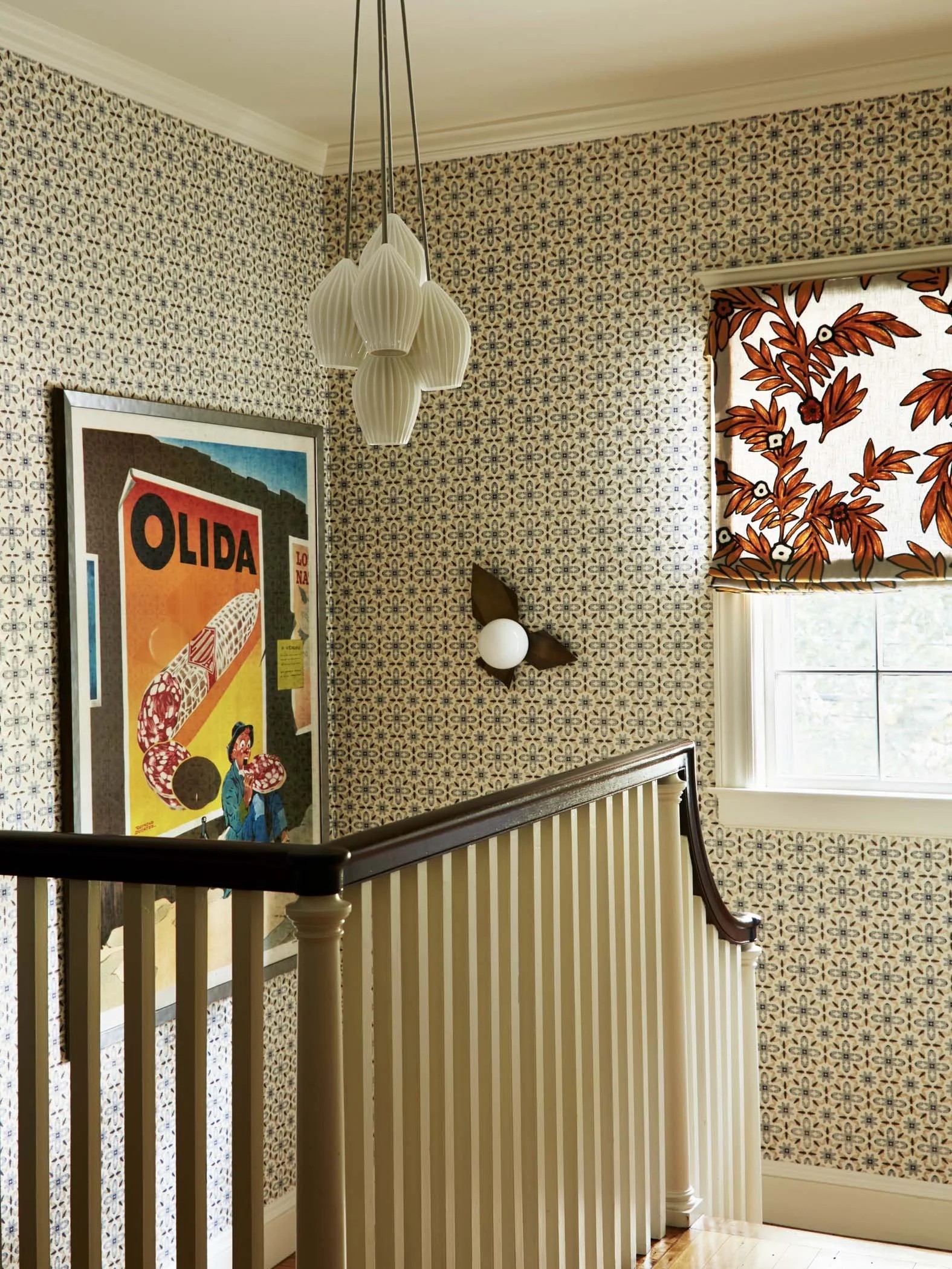 Image of a stairwell with beige and blue patterned wallpaper and a window with a curtain that is white with large orange flowers. There is a large print hanging on the wall and a white light fixture overhead.