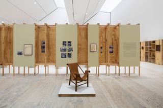 The ’Lina Bo Bardi: Habitat’ exhibition at Museo Jumex. A gallery with a wooden leather chair on a square platform in the center of the room in front of a wooden wall partition with photos and paintings on it.
