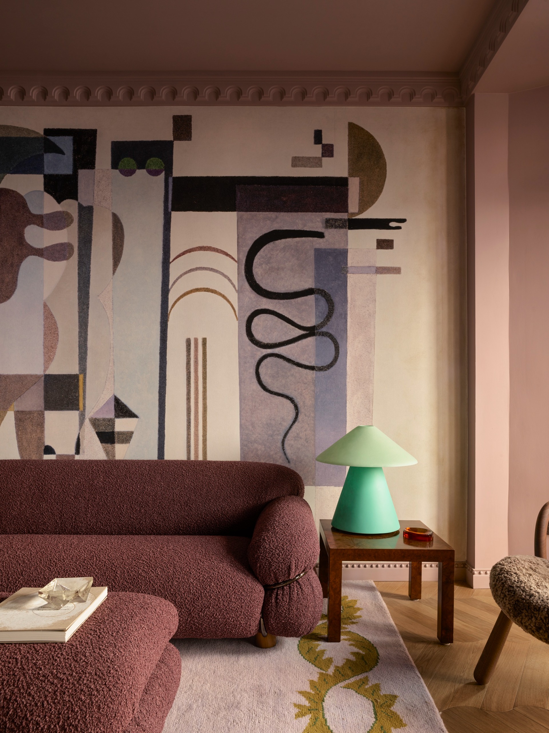 pink living room with wall mural, purple boucle sofa and coffee table, timber side table with mint green lamp, pink rug with yellow border and sheepskin shearling armchair