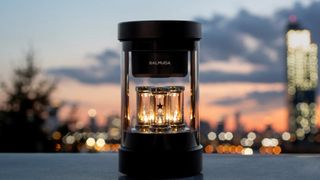 Balmuda the Speaker Bluetooth-Lautsprecher auf einem dunklen Tisch, mit beleuchteten Lichtern und einer Stadtlandschaft im Hintergrund bei Sonnenuntergang