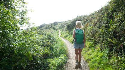 A woman in shorts and hiking boots walks along a rural path. She is facing away from the camera, wearing a backpack, and on either side of the path she walks on are lots of leafy bushes and plants.