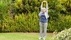 A woman stretches in a garden, holding her hands overhead and interlocking her fingers.