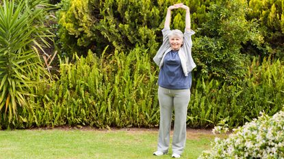 A woman stretches in a garden, holding her hands overhead and interlocking her fingers.