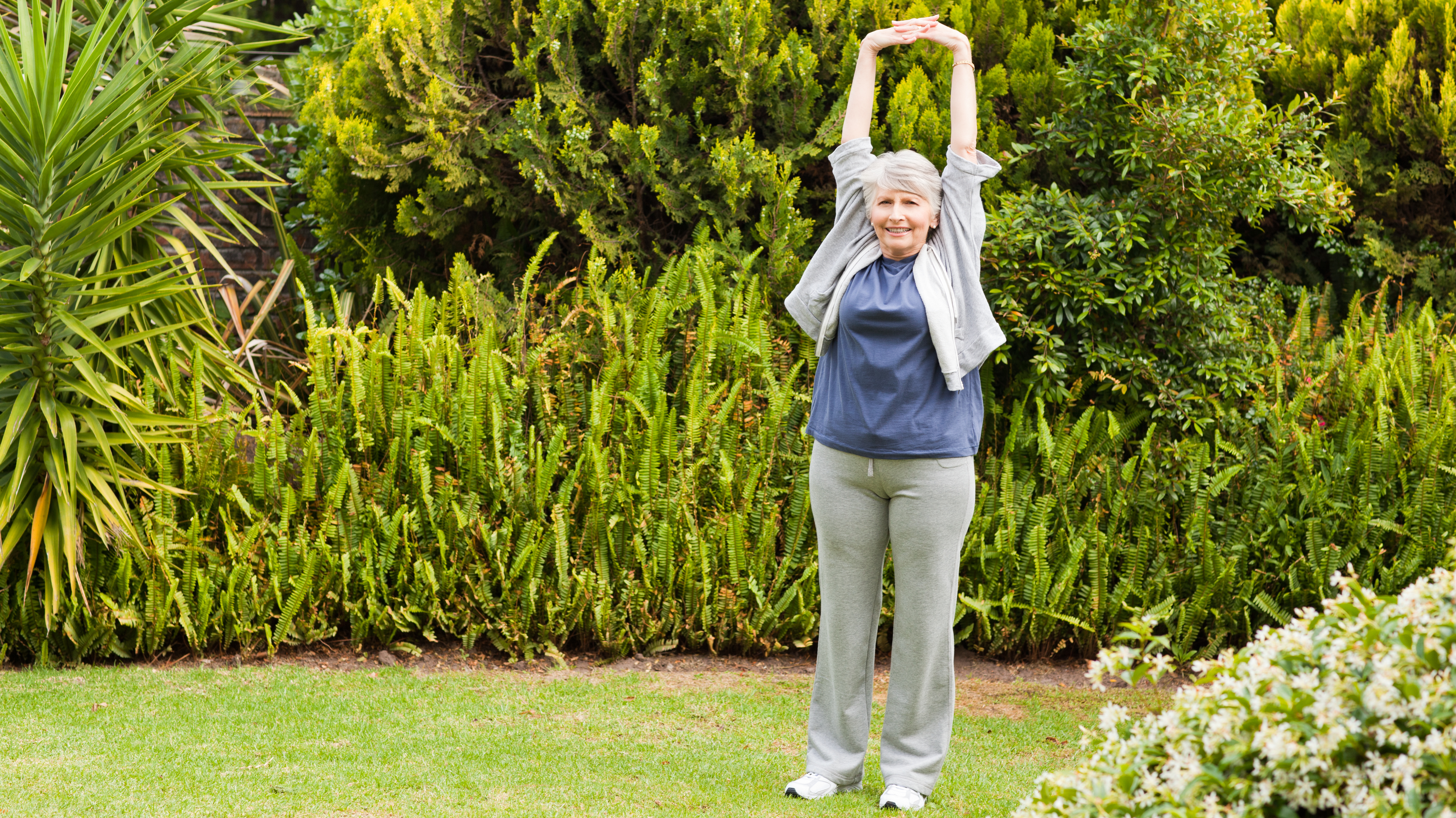 A woman stretches in a garden, holding her hands overhead and interlocking her fingers.