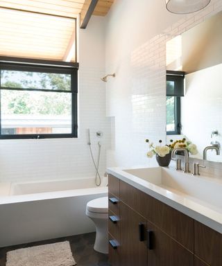 A small bathroom with white subway tiles, a black window, a white bath tub, and a dark brown wooden sink unit with a large rectangular mirror above it