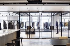 Interior view of Dunhill’s HQ featuring a light coloured counter, black bar stools, a wooden table and chairs and a showroom with glass partitions, glass doors and rails of clothing inside plus a mannequin displaying a suit