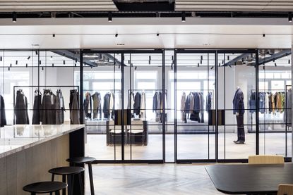 Interior view of Dunhill’s HQ featuring a light coloured counter, black bar stools, a wooden table and chairs and a showroom with glass partitions, glass doors and rails of clothing inside plus a mannequin displaying a suit