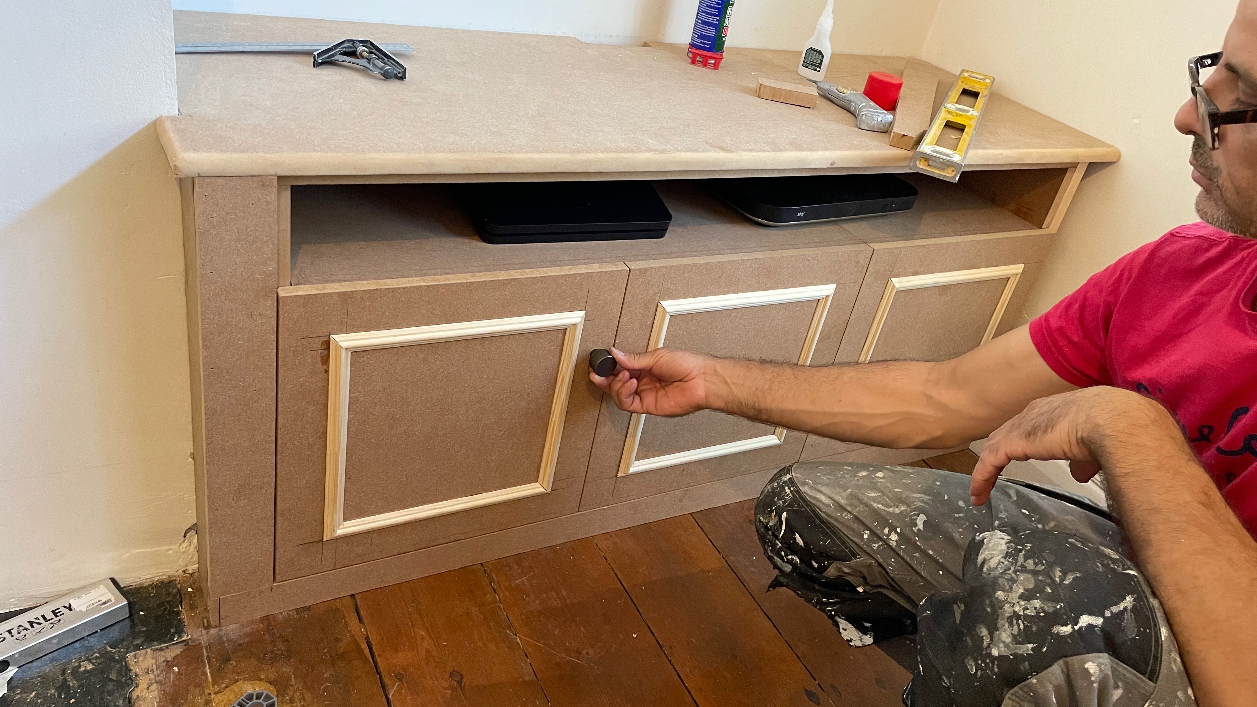 A carpenter holding a door handle in place on an unfinished TV cabinet.