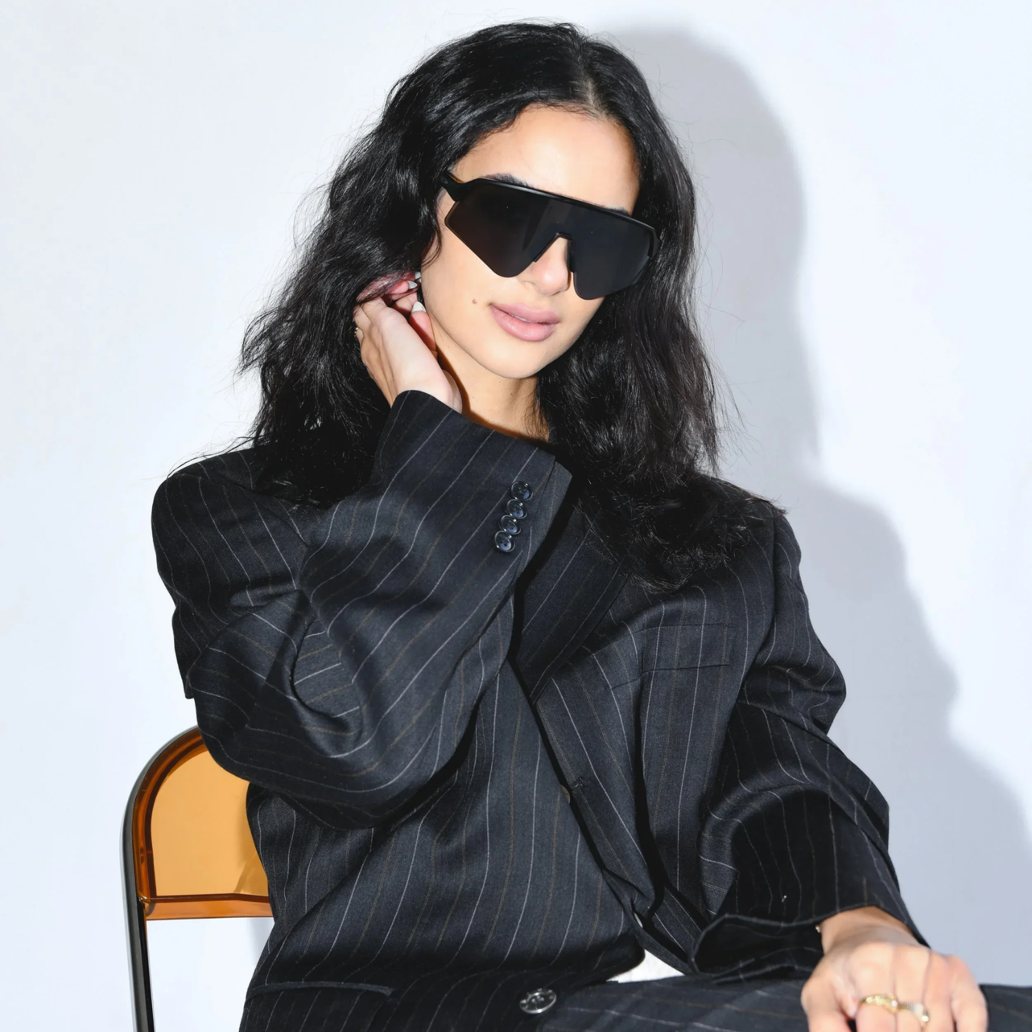 A headshot of a woman with long black hair wearing a pair of oversized sport sunglasses in a pinstripe black blazer