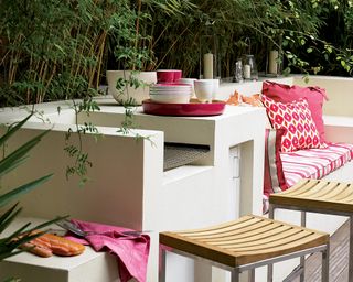 An example of colorful outdoor kitchen designs with white painted stone seating and kitchen units and bright pink and orange pillows, textiles and crockery.