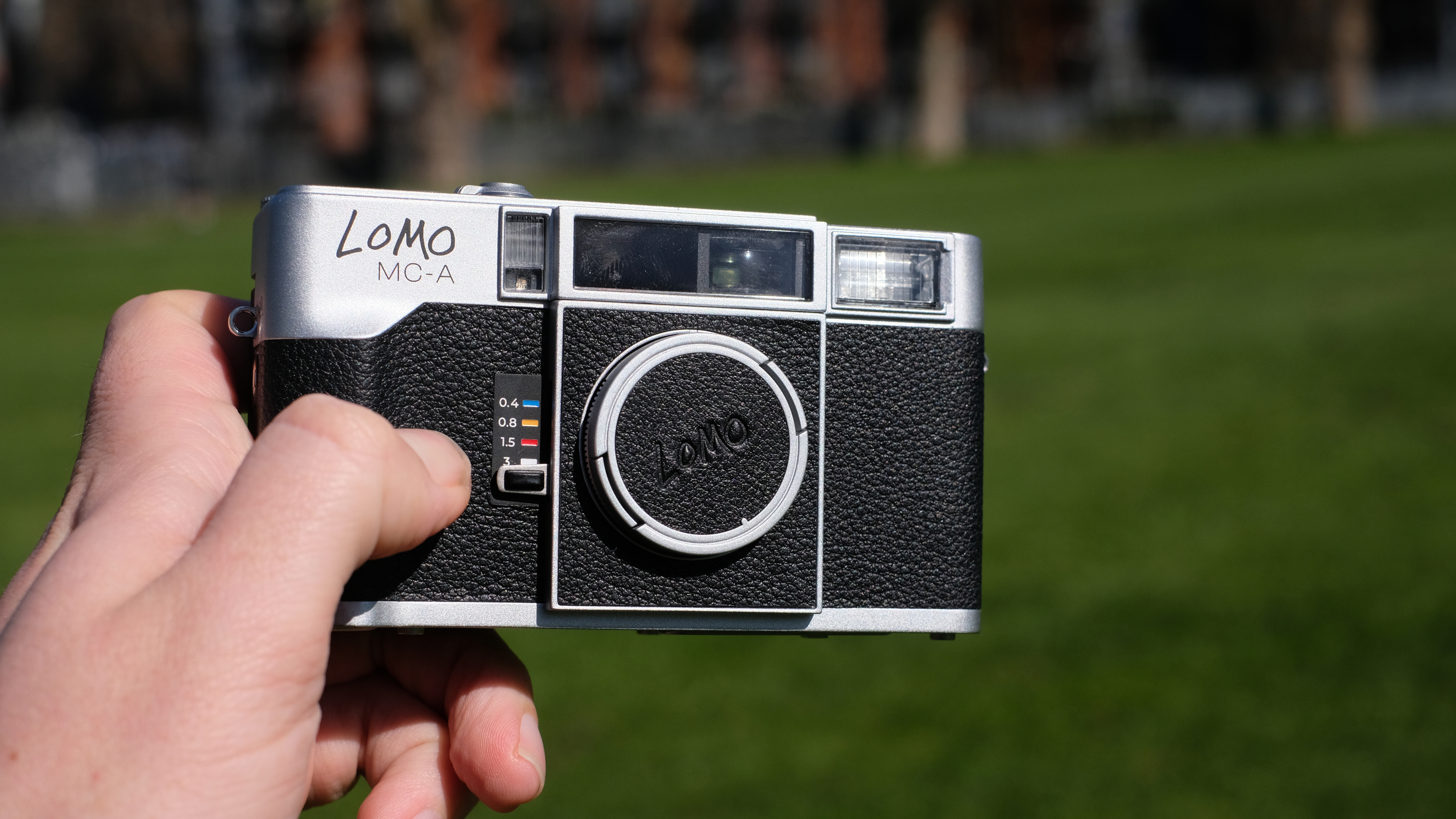 Man's hand holding the Lomography Lomo MC-A analog compact camera, front-facing, with lens cap on