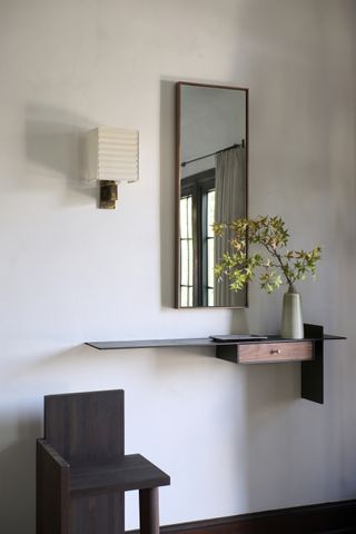 Image of a white wall with a floating shelf with a small drawer hung in the middle. There is a rectangular mirror above the shelf and a small sconce light with a paper lantern shade. There is also a small wooden chairs to the left of the shelf.