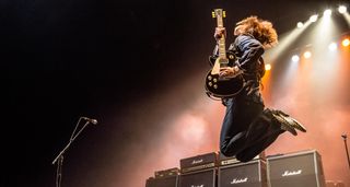 A guitarist with long curly hair and a Les Paul jumps onstage in front of a wall of Marshall amps