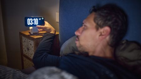 A man with short brown hair wakes up in the middle of the night and makes the sleep hygiene mistake of looking at his alarm clock, which shows the time at 3.10am.
